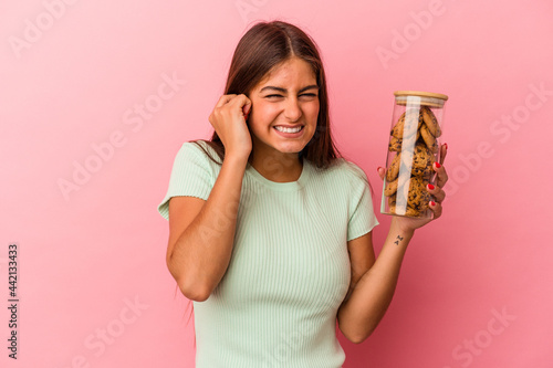 Young caucasian woman holding a cookies jar isolated on pink background covering ears with hands.