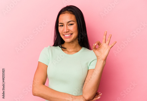 Young Venezuelan woman isolated on pink background winks an eye and holds an okay gesture with hand. © Asier