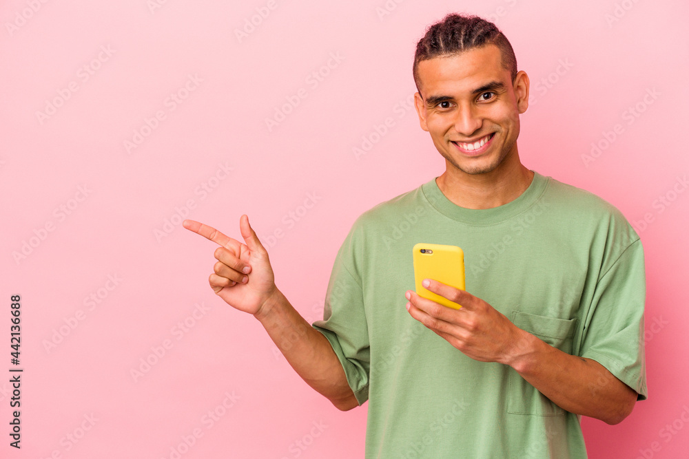 Young Venezuelan man holding a mobile phone isolated on pink background smiling and pointing aside, showing something at blank space. Young Venezuelan man holding a mobile phone isolated on pink background smiling and pointing aside, showing something at blank space.