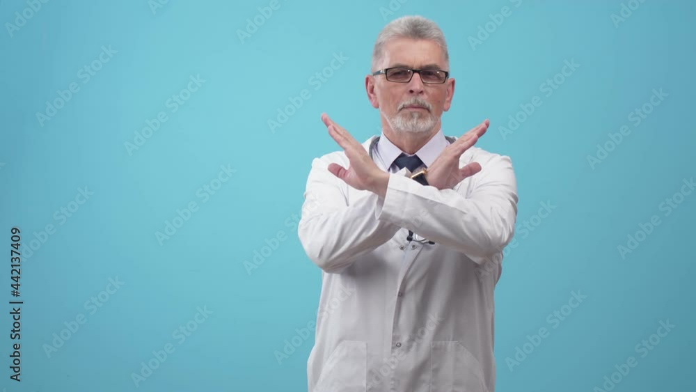 Doctor a man in medical form, shows the gesture of cross with hands and negatively moves head, looking at camera on the blue background of studio. Doctor a man in medical form, shows the gesture of cross with hands and negatively moves head, looking at camera on the blue background of studio.