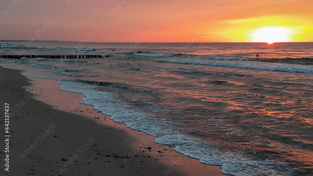 Szenischer Strand mit Wasserwellen und orangenem Sonnenuntergang. Die Sonne spiegelt sich orange bis pink auf dem im flachen Winkel fotografiertem Wasser.