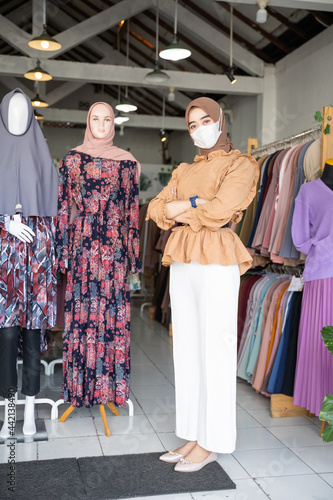 a woman in a headscarf wearing a mask and crossing her arms stands inside a boutique shop