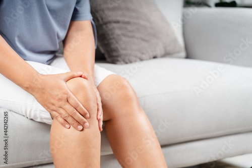 Close up of chubby woman sitting on the sofa and feeling knee pain and she massages her knee. Healthcare and medical concept..