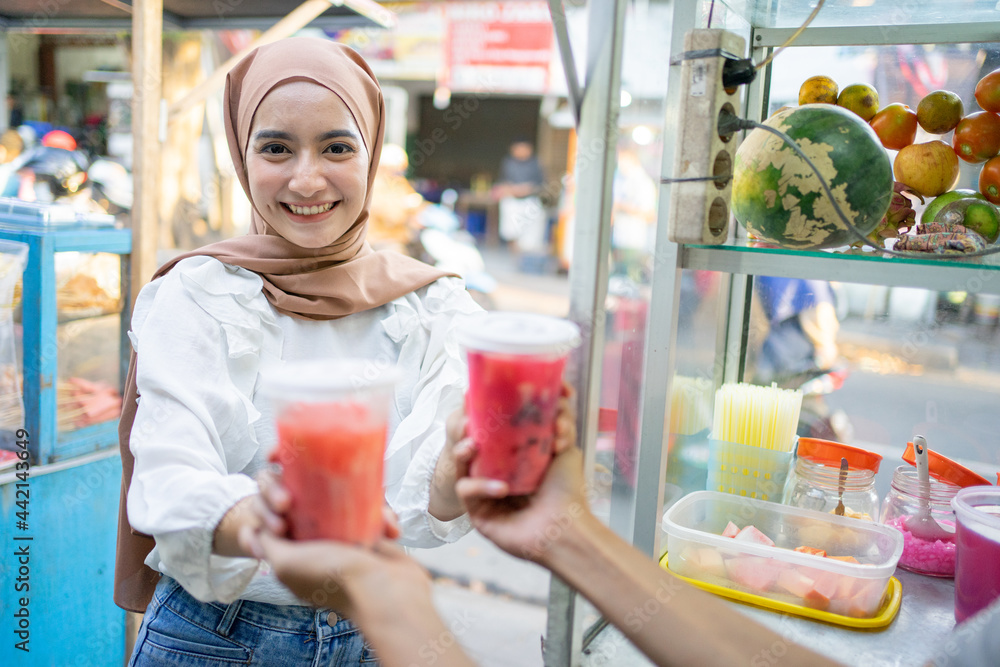 Fototapeta premium a pretty girl in a head scarf smiles at the camera as she picks up two cups of fruit ice from the seller beside the cart