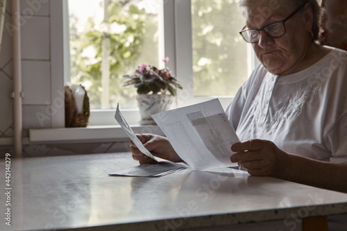 Elderly woman sitting at table in kitchen at home holds domestic bills, feels concerned forgot to pay or debt formed. Middle aged 80s retired woman