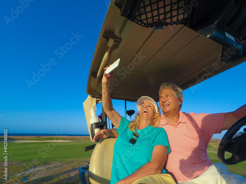 Active senior couple playing golf at sunset sitting on the golf cart with cell phone.