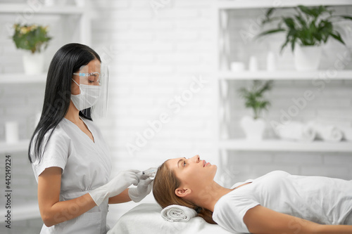 Side view of beautician in white rubber gloves holding injection and doing special procedure for improvements hair for young brunette woman. Concept of beauty procedure in professional salon. 