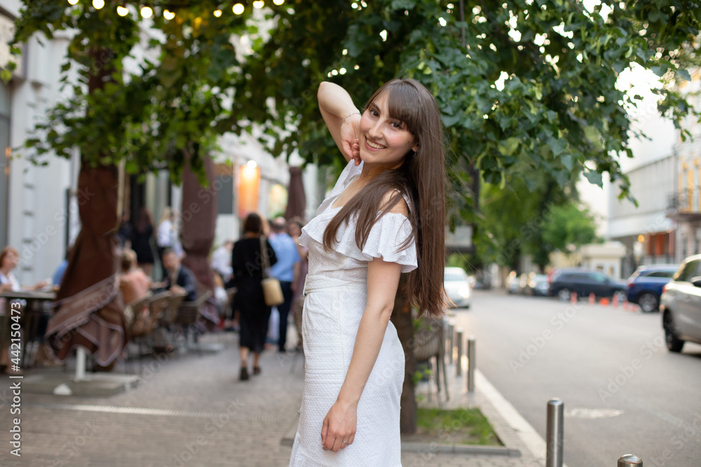 smiling brunette girl with long hair in white dress on background of green trees in the city
