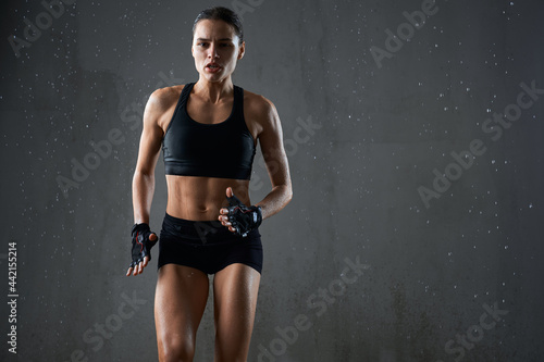 Portrait of wet female bodybuilder with perfect muscular body jogging on spot isolated in empty hall, loft interior. Concentrated athletic woman practicing hard cardio sport exercise under rain.