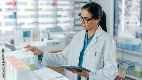 Fotografie Pharmacy Drugstore: Beautiful Caucasian Pharmacist Uses Digital Tablet Computer, Checks Inventory of Medicine, Drugs, Vitamins, Health Care Products on a Shelf