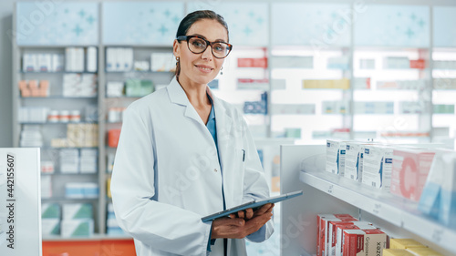 Obraz na plátně Pharmacy: Portrait of Professional Beautiful Caucasian Female Pharmacist Uses Digital Tablet Computer, Checks Medicine Inventory, Looks at Camera, Smiles Charmingly