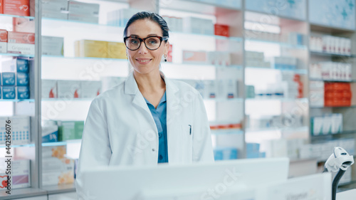 Pharmacy Counter  Portrait of Beautiful Professional Caucasian Female Pharmacist Working on Computer  Looks at Camera Smiling Charmingly. Drugstore Store with Shelves Health Care Products