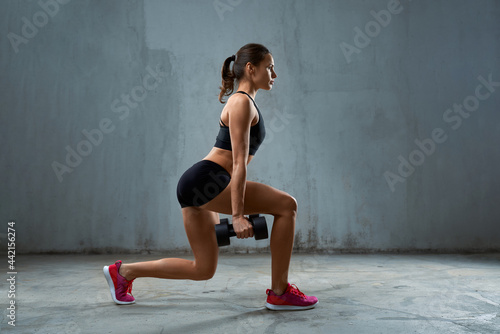 Side view of female bodybuilder training legs using dumbbells. Concentrated fit sportswoman with muscular body doing lunges in empty gym, loft interior. Concept of bodybuilding, healthy lifestyle.