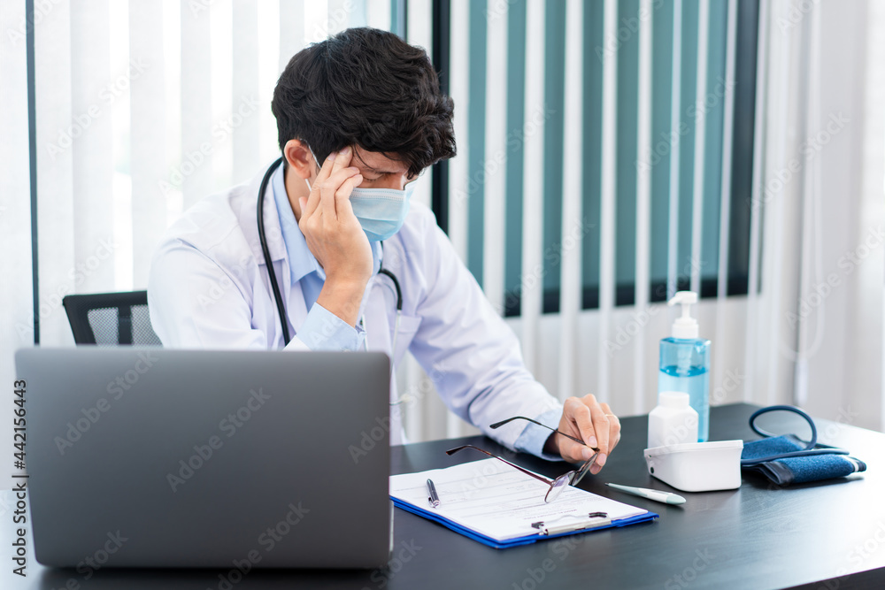 Medical concept a male doctor wearing a mask having a headache while his hard working caused by for Coronavirus outbreak (COVID-19) at his modern clinic Medical concept a male doctor wearing a mask having a headache while his hard working caused by for Coronavirus outbreak (COVID-19) at his modern clinic