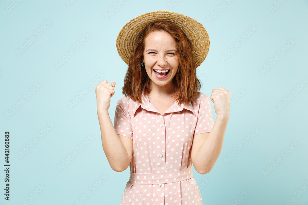 Excited jubilant overjoyed happy young redhead curly woman in casual pink dress straw hat doing winner gesture celebrate clench fists say yes isolated on pastel blue color background studio portrait.