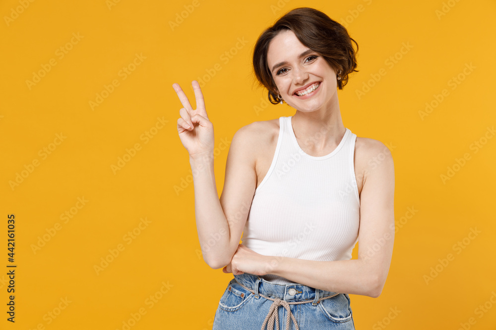 Young cheerful friendly smiling happy woman 20s with bob haircut in white tank top shirt show victory v-sign gesture look camera isolated on yellow color background studio People lifestyle concept Young cheerful friendly smiling happy woman 20s with bob haircut in white tank top shirt show victory v-sign gesture look camera isolated on yellow color background studio People lifestyle concept