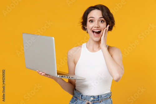 Young freelancer surprised happy woman 20s with bob haircut wearing white tank top shirt using laptop pc computer chat online browsing surfing internet hold face isolated on yellow color background. photo