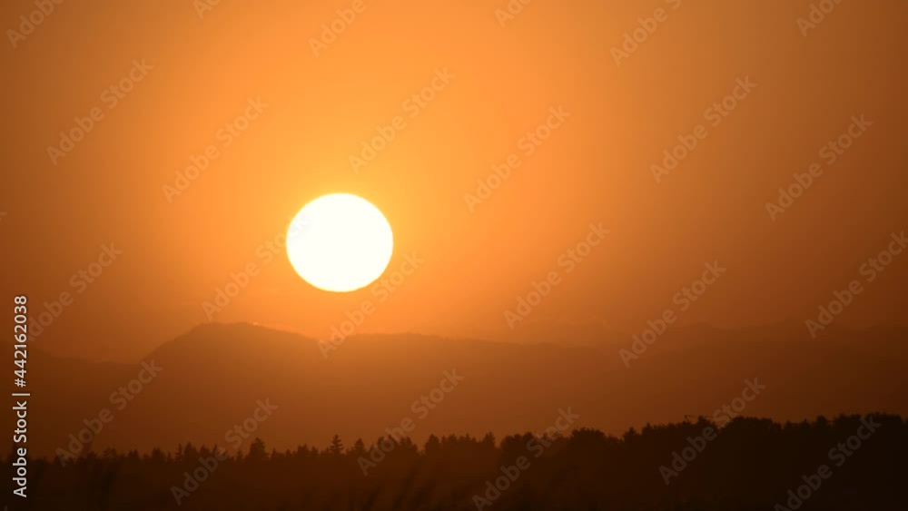 Sun setting in orange sky. Big bright yellow sun moves down. Silhouette of forest and mountains on the horizon. Amazing golden hour colors. Beautiful sunset. Real time, long lens Sun setting in orange sky. Big bright yellow sun moves down. Silhouette of forest and mountains on the horizon. Amazing golden hour colors. Beautiful sunset. Real time, long lens