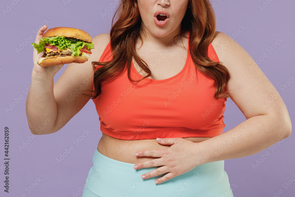 Close up cropped photo young chubby overweight plus size fit woman in red top warm up training hold fastfood burger put hand belly isolated on purple background gym Workout sport motivation concept.