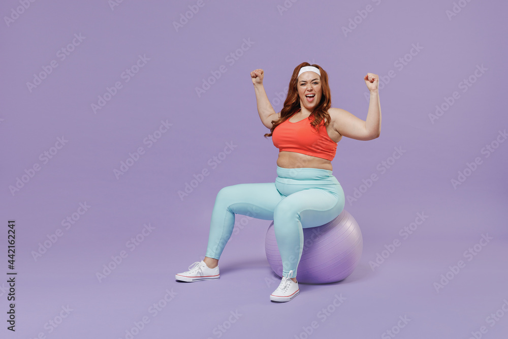 Full length side view young chubby overweight plus size big fat fit woman in red top warm up train sit on fit ball do winner gesture isolated on purple background gym Workout sport motivation concept