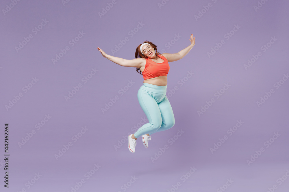 Full length young chubby overweight plus size big fat fit woman wear red top warm up train jump high with outstretched hands isolated on purple background home gym. Workout sport motivation concept