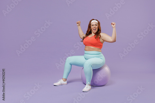 Full length side view young chubby overweight plus size big fat fit woman in red top warm up train sit on fit ball do winner gesture isolated on purple background gym Workout sport motivation concept © ViDi Studio