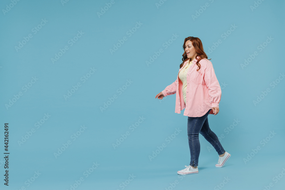 Full length side view young fun smiling redhead chubby overweight woman 30s wearing in pink shirt jeans casual clothes walking going looking aside isolated on pastel blue background studio portrait Full length side view young fun smiling redhead chubby overweight woman 30s wearing in pink shirt jeans casual clothes walking going looking aside isolated on pastel blue background studio portrait