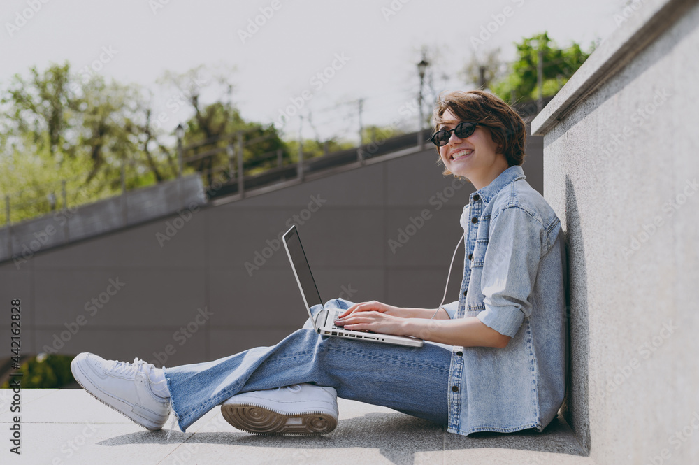 Side view full length young freelancer smiling fun student woman in jeans clothes headphones eyeglasses listen to music leaning on building wall sit on concrete steps outdoors use laptop pc computer.