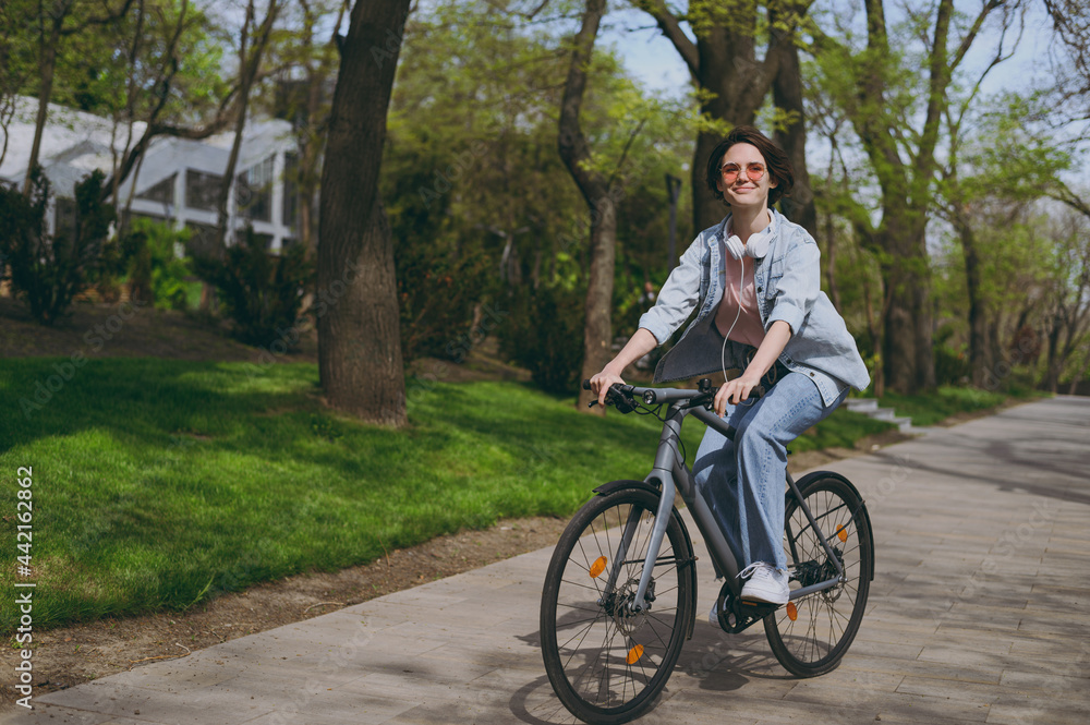 Full length side view young sporty woman in jeans clothes headphones riding bicycle bike on sidewalk in city spring park outdoors, look aside. People active urban healthy lifestyle cycling concept