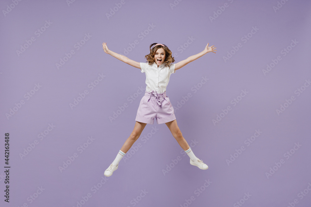 Full length little fun overjoyed kid girl 12-13 years old in white short sleeve shirt jumping high with outstretched hands isolated on purple color background. Childhood children lifestyle concept