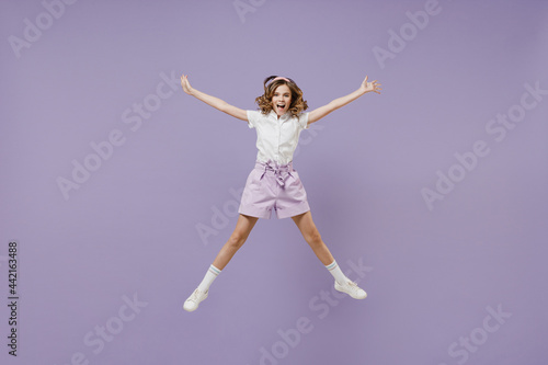 Full length little fun overjoyed kid girl 12-13 years old in white short sleeve shirt jumping high with outstretched hands isolated on purple color background. Childhood children lifestyle concept