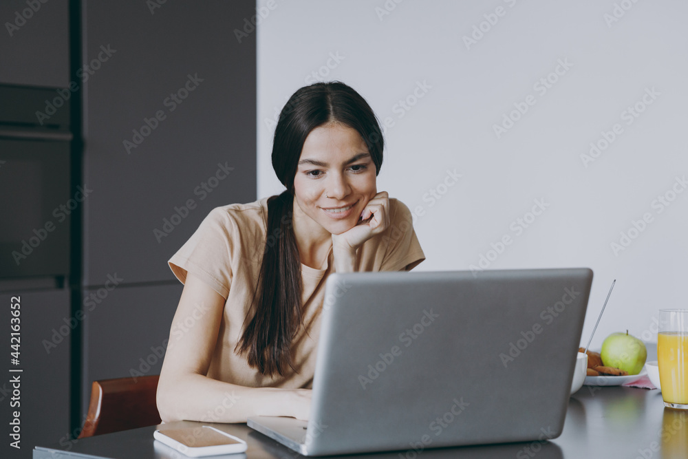 Young happy freelenacer copywriter housewife woman 20s in beige shirt eat breakfast using laptop pc computer look for recipe cook food in light kitchen at home alone. Healthy diet lifestyle concept. Young happy freelenacer copywriter housewife woman 20s in beige shirt eat breakfast using laptop pc computer look for recipe cook food in light kitchen at home alone. Healthy diet lifestyle concept.