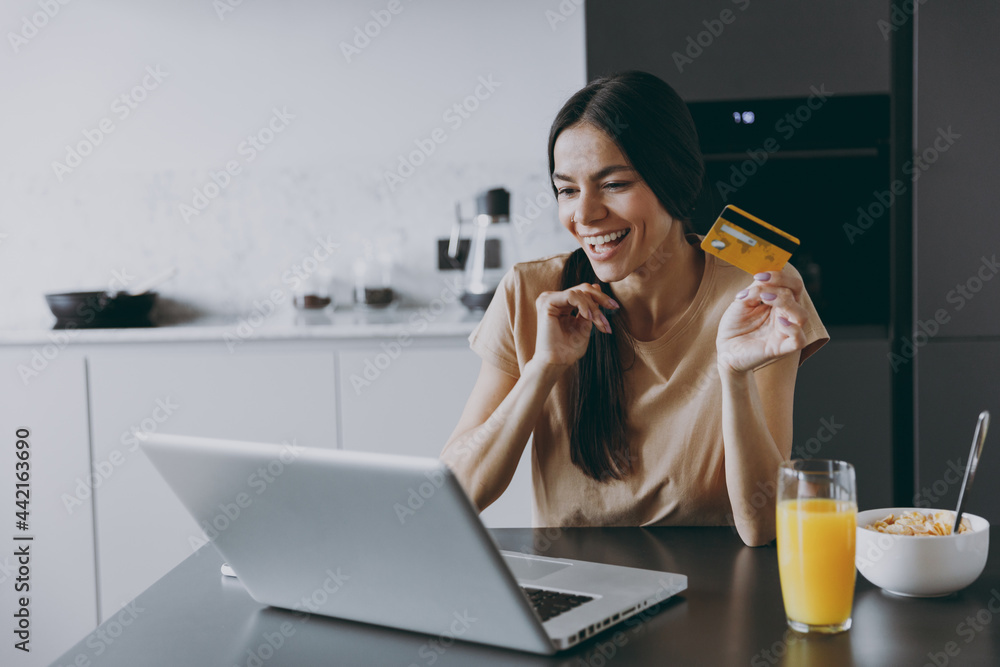 Young happy housewife woman 20s in shirt eat breakfast use laptop pc computer credit bank card shopping online order delivery cook food in light kitchen at home alone Healthy diet lifestyle concept. Young happy housewife woman 20s in shirt eat breakfast use laptop pc computer credit bank card shopping online order delivery cook food in light kitchen at home alone Healthy diet lifestyle concept.