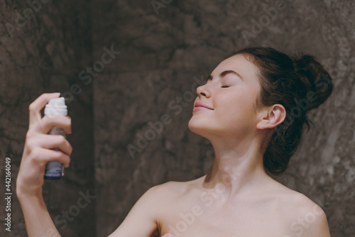 Side view half naked topless young satisfied woman 20s apply on fixative and make-up base on face with closed eye in bathroom brown dark background. Skin care healthcare cosmetic procedures concept