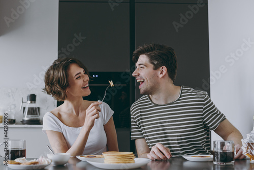 Young happy couple two woman man 20s in casual t-shirt clothes sit by table eat pancakes with maple syrup feed husband cooking food in light kitchen at home together Healthy diet lifestyle concept