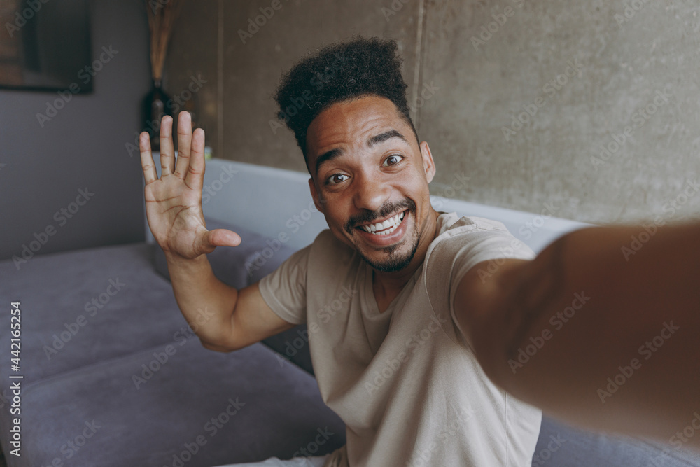 Close up fun happy smiling young african american man in beige casual t-shirt sit on grey sofa indoors apartment doing selfie shot on mobile phone waving hand say hello, rest on weekends stay at home - obrazy, fototapety, plakaty 