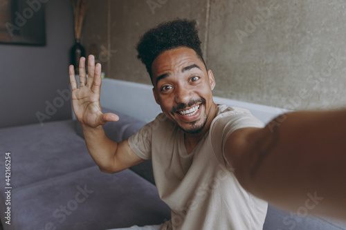 Close up fun happy smiling young african american man in beige casual t-shirt sit on grey sofa indoors apartment doing selfie shot on mobile phone waving hand say hello, rest on weekends stay at home