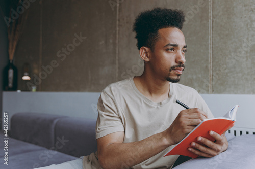 Pensive thoughtful young poet student african american man 20s wearing beige t-shirt sitting on grey sofa indoors apartment write down memories in notebook diary, resting on weekends staying at home photo