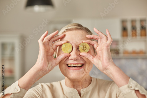 An elderly woman holds bitcoin coins in her hands near her eyes. Seniors retirement happy life concept