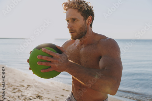 Young sporty athletic toned fit sportsman topless man 20s warm up train play hold medicine crossfit ball at sunrise sun dawn over sea beach outdoor on sand dawn beach seaside in summer day morning. photo