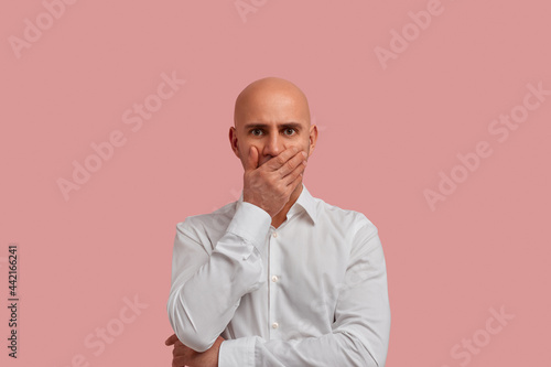 I dont want to believe it. Disappointed bald man with bristle, covered mouth with hand, shocked information about his friend. Dressed in white shirt, isolated over pink background.
