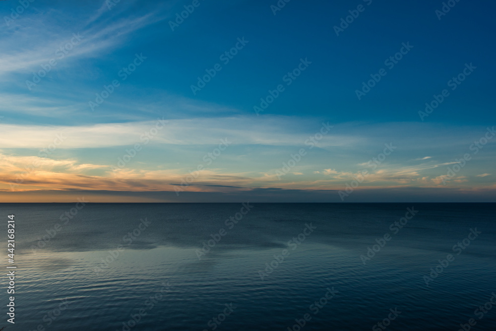 A sky with haze turning into cloudy on the horizon, during the white nights at sunset over a calm sea with fine ripples.