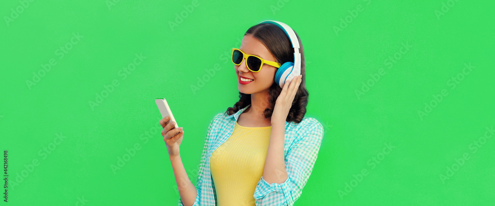 Portrait of modern happy smiling young woman looking at smartphone listening to music in wireless headphones on green background