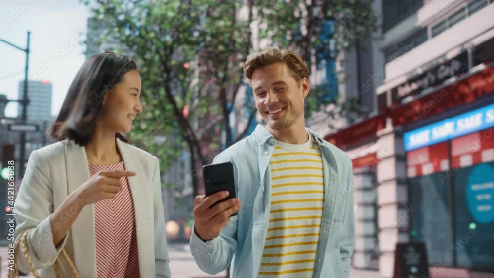 Young Stylish Multiethnic Couple is Casually Walking on a Street in a Big City. Handsome Caucasian Male Showing Smartphone to Beautiful Japanese Female. Diverse Friends Enjoying Travelling Together. Young Stylish Multiethnic Couple is Casually Walking on a Street in a Big City. Handsome Caucasian Male Showing Smartphone to Beautiful Japanese Female. Diverse Friends Enjoying Travelling Together.