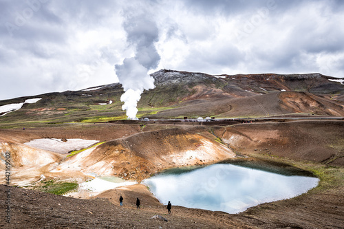 People walking hiking on Viti Crater of Krafla caldera geothermal formation with small lake water and mountains snow during summer and geyser hot spring steam photo