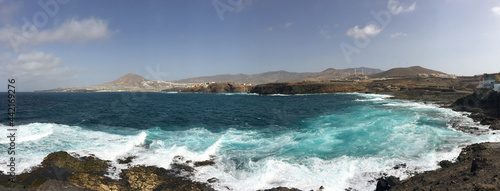 Panoramic photo of the north coast of Gran Canaria with the rough sea with white foam and turquoise and navy blue colors. Gáldar mountain on the horizon under a blue sky. © TravelmeSoftly