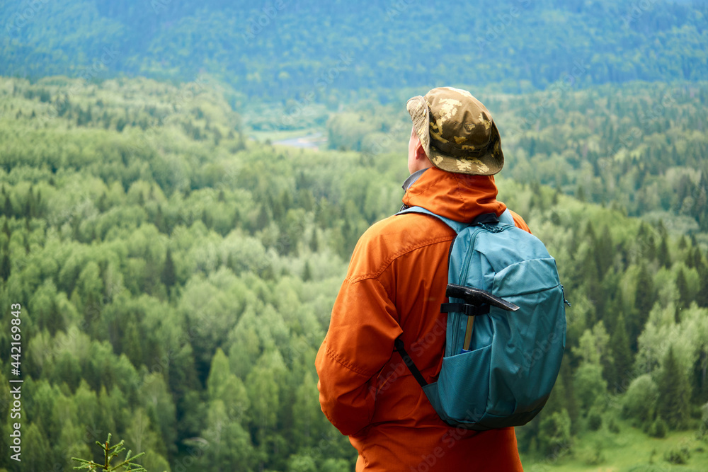 man traveler with a backpack looks down from the mountain at the wooded valley below him