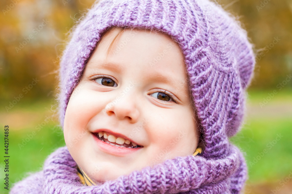 A child in a purple knitted hat and scarf. Portrait of a child against the background of autumn trees. Cute smiling boy. A child in a purple knitted hat and scarf. Portrait of a child against the background of autumn trees. Cute smiling boy.