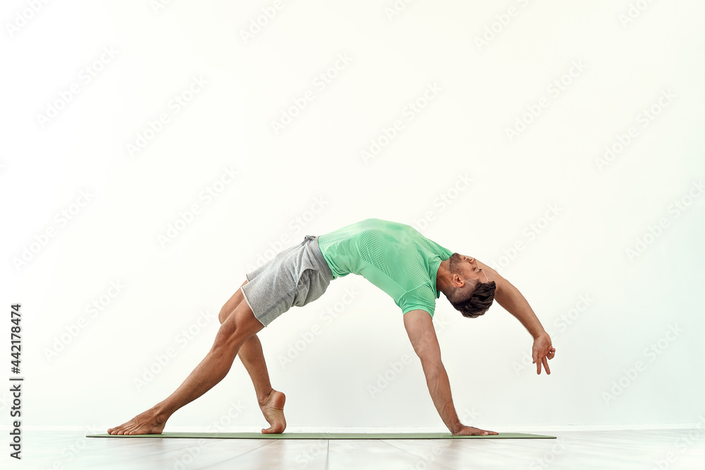 Fit male practicing yoga Wild Thing Pose, Camatkarasana against white wall background