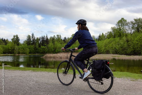 Adult Caucasian Woman Riding a Bicycle on a path by a lake in a modern city park. Spring Evening. Taken in Green Timbers Urban Forest, Surrey, Vancouver, British Columbia, Canada.